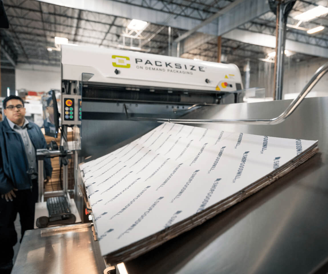 A photo of a Shop Goodwill employee operating a Packsize automated box making machine.